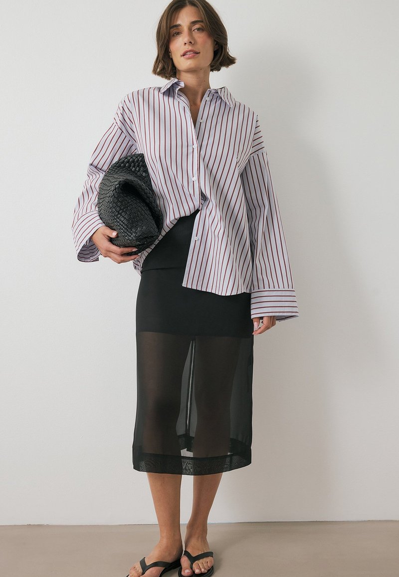 Chemise à rayures blanche et bordeaux. Jupe noire transparente superposée sur une base noire solide. Pochette tissée noire et sandales.