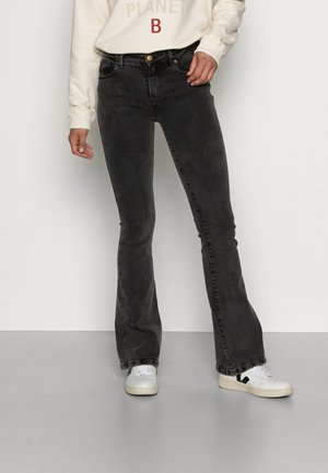 Osoba nosząca wyblakłe czarne jeansy bootcut, białe trampki z czarnym logo oraz kremową bluzę z napisem „PLANET B”.