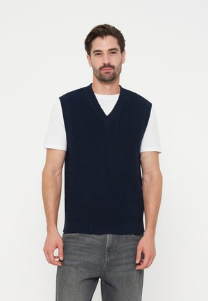 Homme portant un pull sans manches en maille bleu marine à col en V par-dessus un t-shirt blanc et un jean gris, debout devant un fond blanc uni.