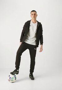 Mladý muž v černém sportovním oblečení Puma a kopačkách stojící s jednou nohou na vícoloré fotbalovém míči na jednobarevném bílém pozadí.