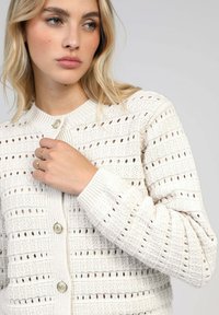 Cardigan en tricot couleur blanc cassé avec un motif ajouré. Il dispose d'un col rond, de poignets côtelés et de deux boutons sur le devant pour la fermeture.