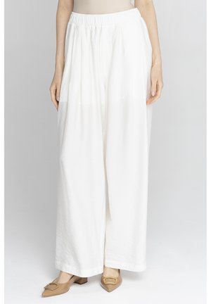 Kobieta nosząca białe, białe spodnie z wysokim stanem i szerokimi nogawkami, w połączeniu z beżowymi niskimi butami na obcasie z złotymi klamrami, stojąca na jednolitym tle.