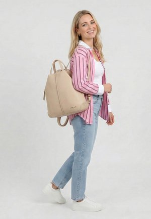 Lachende vrouw die een roze gestreept shirt, wit topje, lichtblauwe spijkerbroek, witte sneakers draagt en een beige leren rugzak op haar schouder heeft.