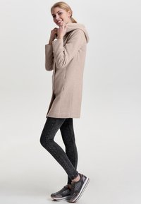 Beige hettejakke med teksturert overflate, brukt over mørke skinny jeans og grå mønstrede sneakers. Jakken har en skreddersydd passform og sidelommer.