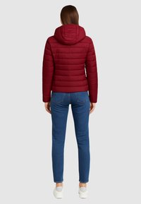 Červená prešívaná puffová bunda s kapucňou, s textúrovaným povrchom a priliehavým strihom, skombinovaná s modrými džínsami a bielymi slip-on topánkami.