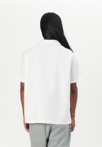 Camicia bianca a maniche corte realizzata in tessuto leggero, con colletto standard, taglio dritto e una texture semplice. Sotto si indossano pantaloni a righe.