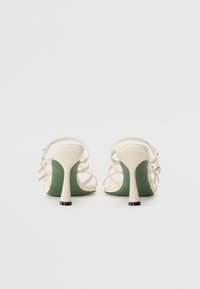 Sandales à talons hauts blanches avec des lanières, finition mate. Accents de talon verts et bases noires. Design à dos ouvert avec une configuration de lanières délicates.