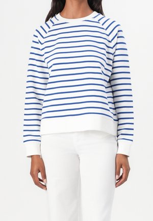 Persona con pantalones blancos y una camisa blanca de manga larga con rayas horizontales azules, de pie frente a un fondo liso.