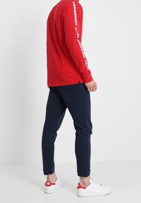 Rode langemouwen shirt met witte tekst op de mouwen, gecombineerd met marineblauwe joggers en witte sneakers met rode accenten, met een eenvoudig ontwerp.