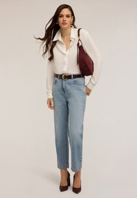 Blouse blanche à boutons, jeans droits bleu clair, ceinture marron avec boucle circulaire, sac à main rouge foncé et escarpins bordeaux.