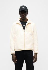 Άνδρας που φοράει εκρού φλις ζακέτα με φερμουάρ, μαύρη μπλούζα The North Face, μαύρο παντελόνι και μαύρο καπέλο The North Face μπροστά σε λευκό φόντο.