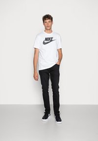 Hvit Nike t-skjorte med svart logo, kombinert med svarte jeans og svarte sneakers. Enfargede; avslappet passform; ingen synlige mønstre eller teksturer.