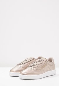Ett par ljusbeige metalliska låga sneakers med vita sulor och snören, placerade sida vid sida på en vit yta.