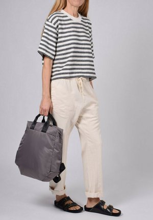 T-shirt courte rayé gris et blanc, pantalon beige avec cordon de serrage, et un sac rembourré gris avec poignées noires. Sandales noires avec clous.