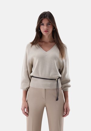 Frau mit langen braunen Haaren, die einen beigen V-Ausschnitt-Pullover mit schwarzem Gürtel und beige Hosen trägt, frontal vor einem weißen Hintergrund.