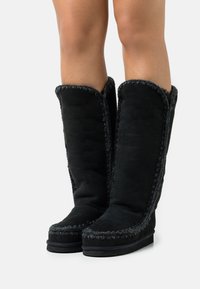 Botas altas de ante negro con costuras texturizadas a lo largo de los bordes yuna suela de goma plana, presentadas en un acabado suave y mate.