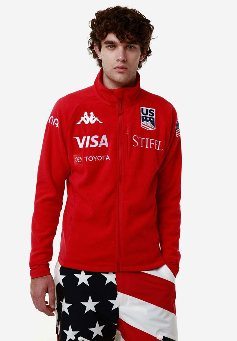 Giacca di pile rossa con colletto alto, con loghi bianchi tra cui "US" e "VISA". Abbinata a pantaloni corti con un motivo a stelle e strisce.