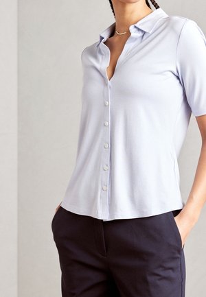 Femme portant une chemise à col boutonnée à manches courtes couleur lavande clair, un pantalon sombre et un simple collier en or, les mains dans les poches.