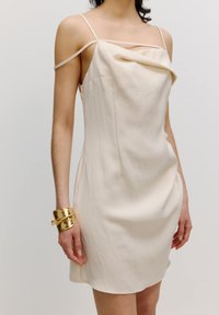 Cremefarbenes, ärmelloses Kleid mit einem Wasserfall-Ausschnitt, aus leichtem Stoff gefertigt. Verfügt über dünne Schulterträger und ein schlichtes Design. Goldfarbener Armreif.