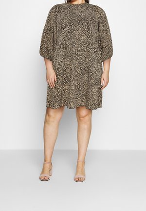 Woman wearing a loose, knee-length, beige and black patterned dress with puffed three-quarter sleeves and beige ankle-strap block heels.