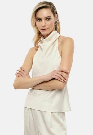 Donna con capelli biondi che indossa un top bianco senza maniche a collo alto e pantaloni bianchi abbinati, braccia incrociate, con orecchini.