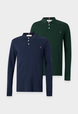 Homme aux cheveux bouclés portant un polo bleu marine à manches longues, un jean bleu clair et des baskets rouges, debout avec une main dans la poche.