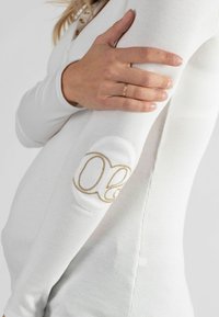 Witte geribde langemouwtop met een gouden geborduurd logo op de elleboog. Het ontwerp omvat een voorste pand met knopen.