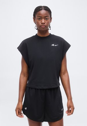 Vrouw draagt een zwarte mouwloze top en zwarte mesh shorts met kleine witte logo’s, staand tegen een effen lichte achtergrond.