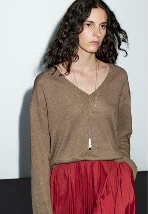 Bruine gebreide trui met een V-hals, gelaagde gouden kettingen, gecombineerd met een rode geplooide rok. Details van de gestructureerde stof zijn zichtbaar.