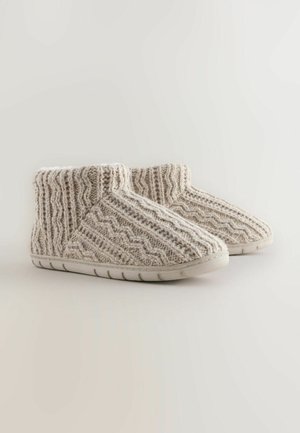 Zapatillas de punto beige texturizadas con forma redondeada, suave forro blanco, suela de goma y un patrón de zigzag distintivo en la tela superior.