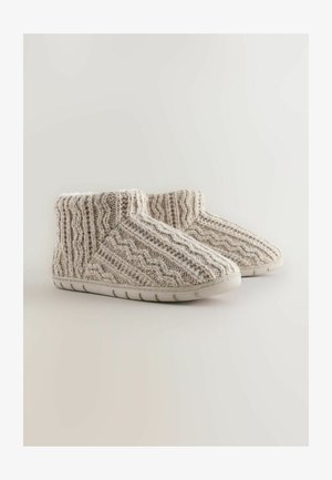 Zapatillas de punto beige texturizadas con forma redondeada, suave forro blanco, suela de goma y un patrón de zigzag distintivo en la tela superior.