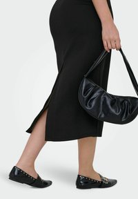 Schwarzes knöchellanges Kleid mit seitlicher Schlitze, kombiniert mit schwarzen flachen Schuhen mit silbernen Nieten und einer schwarzen Handtasche aus Kunstleder mit gerafftem Detail.