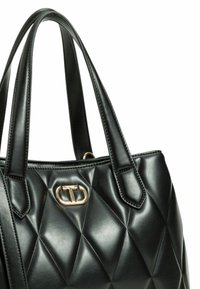 Borsa nera in ecopelle trapuntata con due manici corti, un emblema dorato davanti e dettagli di cucitura elegante.