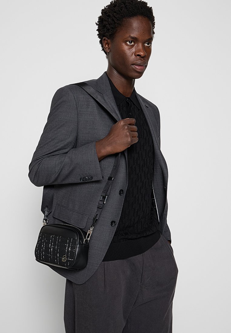Bolso bandolera negro con detalles plateados verticales, forma rectangular y correa ajustable. Llevado sobre un suéter oscuro y una blazer gris.
