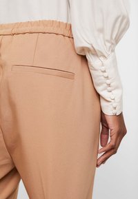 Ljust bruna, byxor med elastisk midja och en slät textur. Bakficka synlig. Vit, texturerad blus med knappdetaljer vid manschetten.