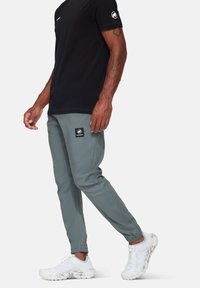 Grijze sportbroek met een elastische tailleband, zijnaad en een logo patch. Draagt met een zwart t-shirt met korte mouwen en witte sneakers.