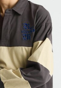 Polohemd mit einem dunkelgrauen Körper und beiger horizontaler Streifen, mit blauem gesticktem Logo auf der Brust und Knopfleiste am Kragen. Weicher Baumwolltextur.