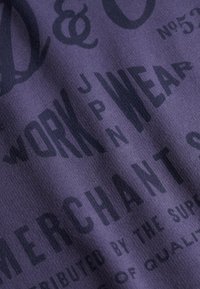 Gros plan sur un tissu violet avec un texte noir comprenant « WORK WEAR » et « MERCHANT » en polices en gras et stylisées.