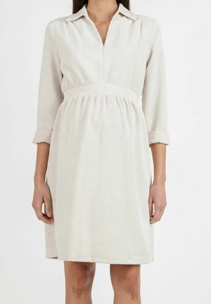 Femme portant une robe en velours côtelé crème longueur genou, avec des manches trois-quarts et une taille froncée.