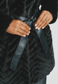 Czarna kurtka z futra syntetycznego o teksturze w zygzaki, z gładkim czarnym skórzanym paskiem związanym w talii, a ręce umieszczone wokół węzła.