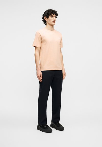 Νεαρός άνδρας με σγουρά μαλλιά που στέκεται, φοράει κοντομάνικη μπλούζα σε ανοιχτό ροδακινί χρώμα, μαύρο παντελόνι και μαύρα αθλητικά παπούτσια σε απλό φόντο.