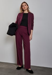 Burgunderfarbener Blazer und passende weite Hosen aus einem weichen Stoff, kombiniert mit einem schwarzen Oberteil. Das Modell hält eine schwarze Steppjacke. Schwarze Schuhe.