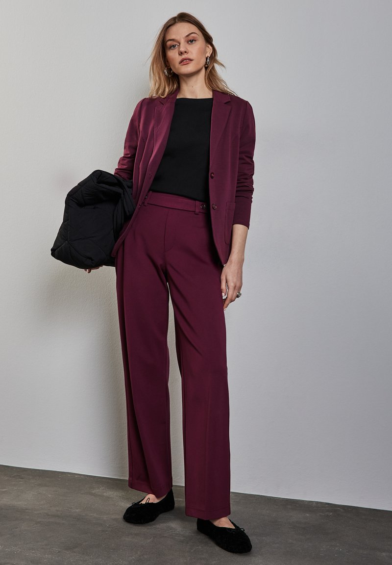 Burgunderfarbener Blazer und passende weite Hosen aus einem weichen Stoff, kombiniert mit einem schwarzen Oberteil. Das Modell hält eine schwarze Steppjacke. Schwarze Schuhe.