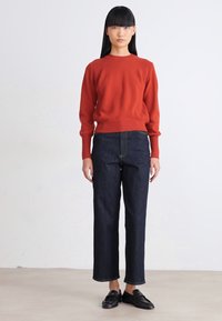 Frau steht in einem enganliegenden rost-orangefarbenen Pullover, dunkelblaue gerade Jeans und schwarze Slipper vor einem einfachen hellen Hintergrund.
