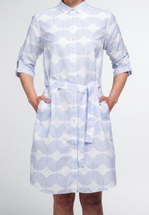Donna che indossa un vestito a bottoni con motivo blu e bianco, maniche arrotolate e cintura abbinata, con le mani nelle tasche, in piedi contro uno sfondo bianco.