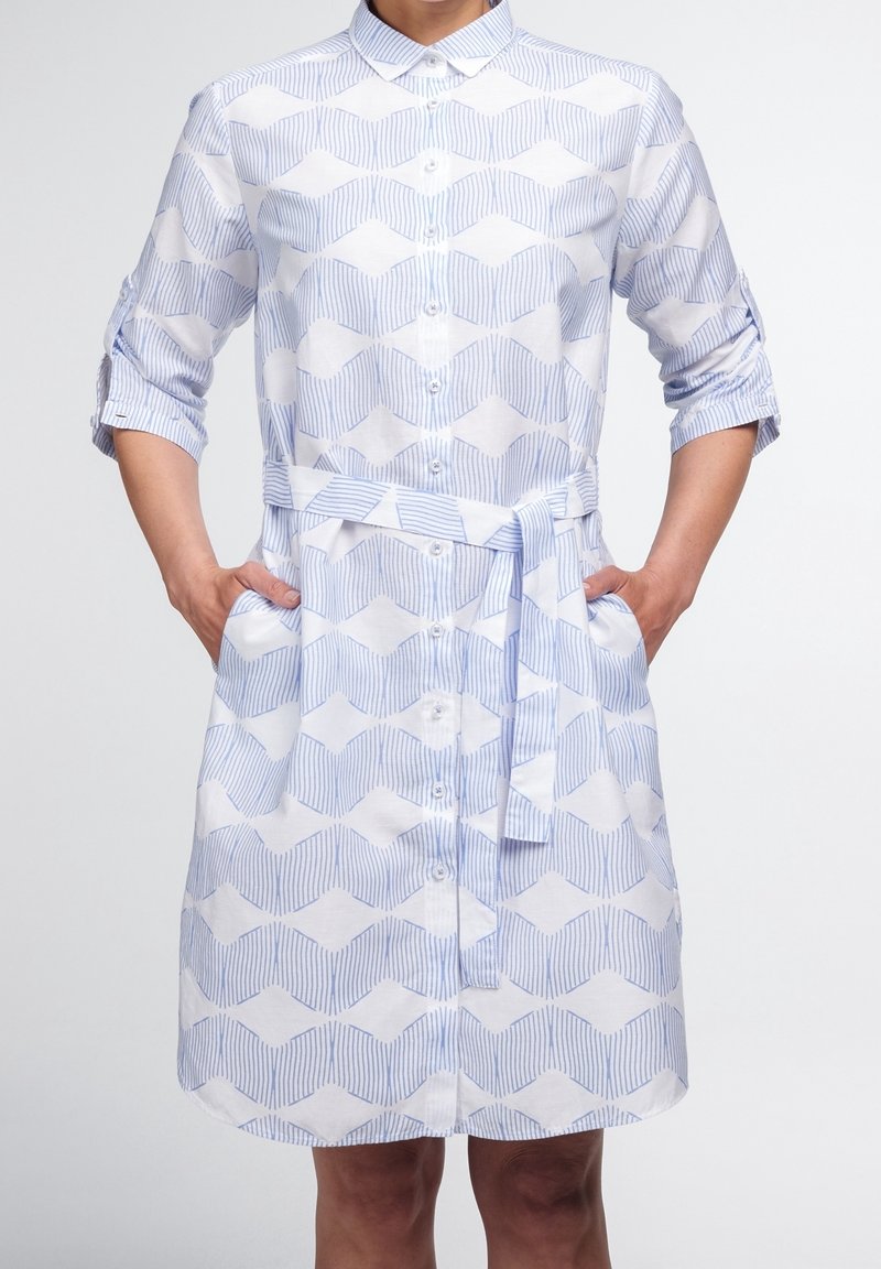 Femme portant une robe à boutons bleue et blanche à motifs, manches retroussées et ceinture assortie, mains dans les poches, debout devant un fond blanc.