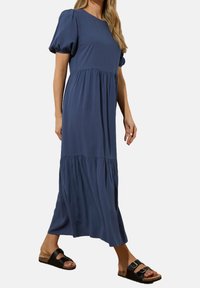 Robe midi bleue avec manches bouffantes et design en couches, avec une texture lisse. Portée avec des sandales noires, affichant un style décontracté.