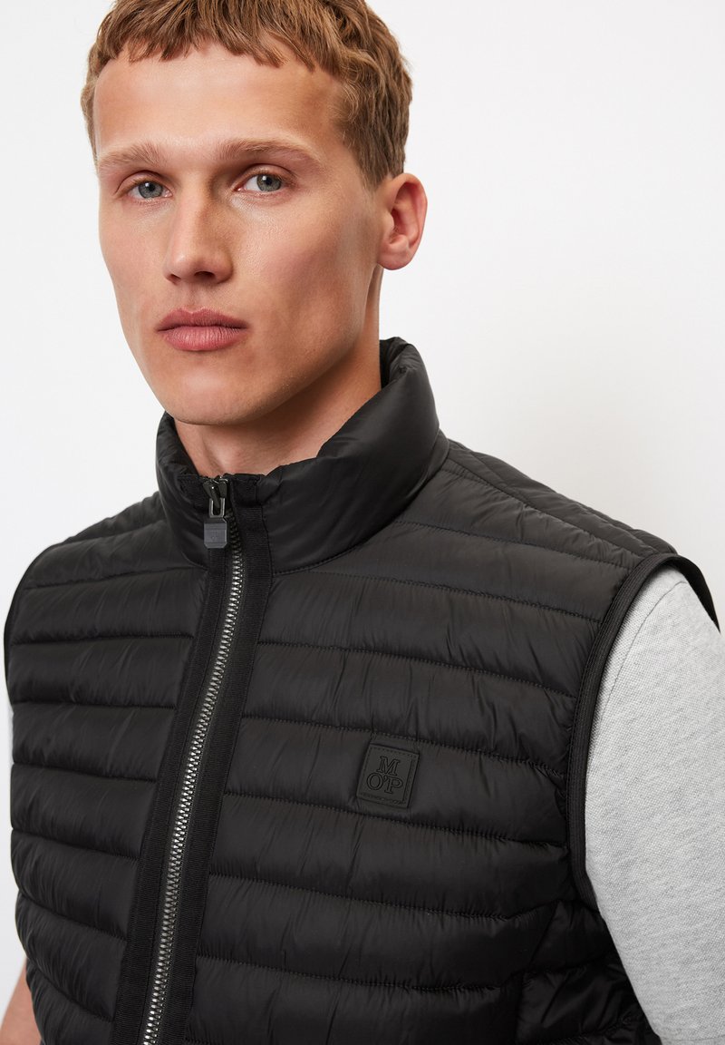 Gilet matelassé noir avec un col montant, fermeture éclair, piquage horizontal et un petit patch logo sur la poitrine. Le tissu semble lisse et léger.