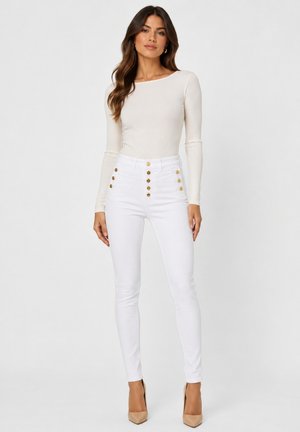Femme aux longs cheveux foncés portant un haut ajusté à manches longues écru, un pantalon blanc taille haute avec des boutons dorés et des talons nus, debout.