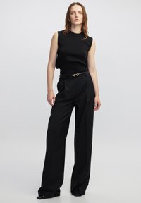 Top senza maniche nero con scollo alto abbinato a pantaloni neri a vita alta. L'outfit presenta una texture liscia e una vestibilità sartoriale, accentuato da una cintura.
